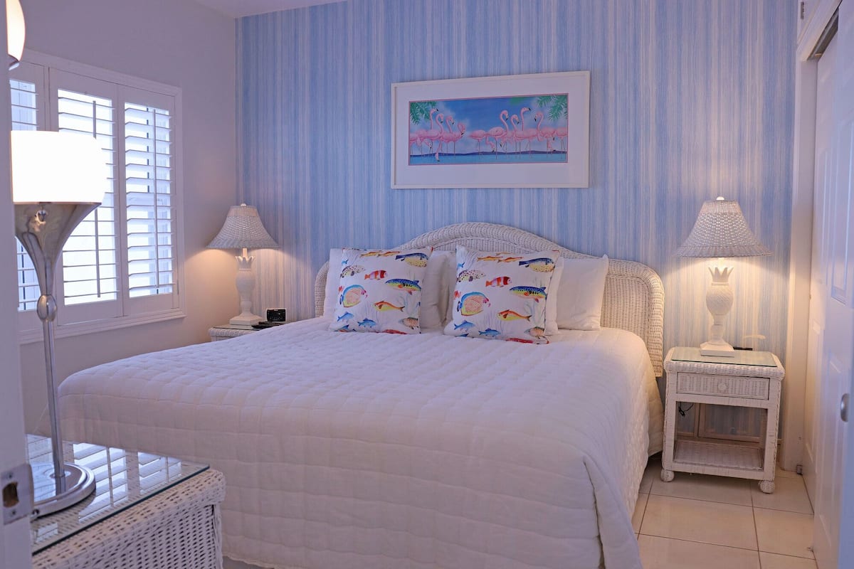A bedroom with a large bed, two bedside tables, a nightstand, a lamp, a framed picture, and a window with blinds.