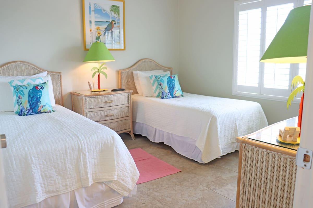 A room with two single beds, a nightstand with a lamp, a small table, and a window with blinds.