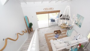 A bright, airy living room with a white sofa, a wooden coffee table, and a chandelier. A sign reading "Beach House" is visible in the background.