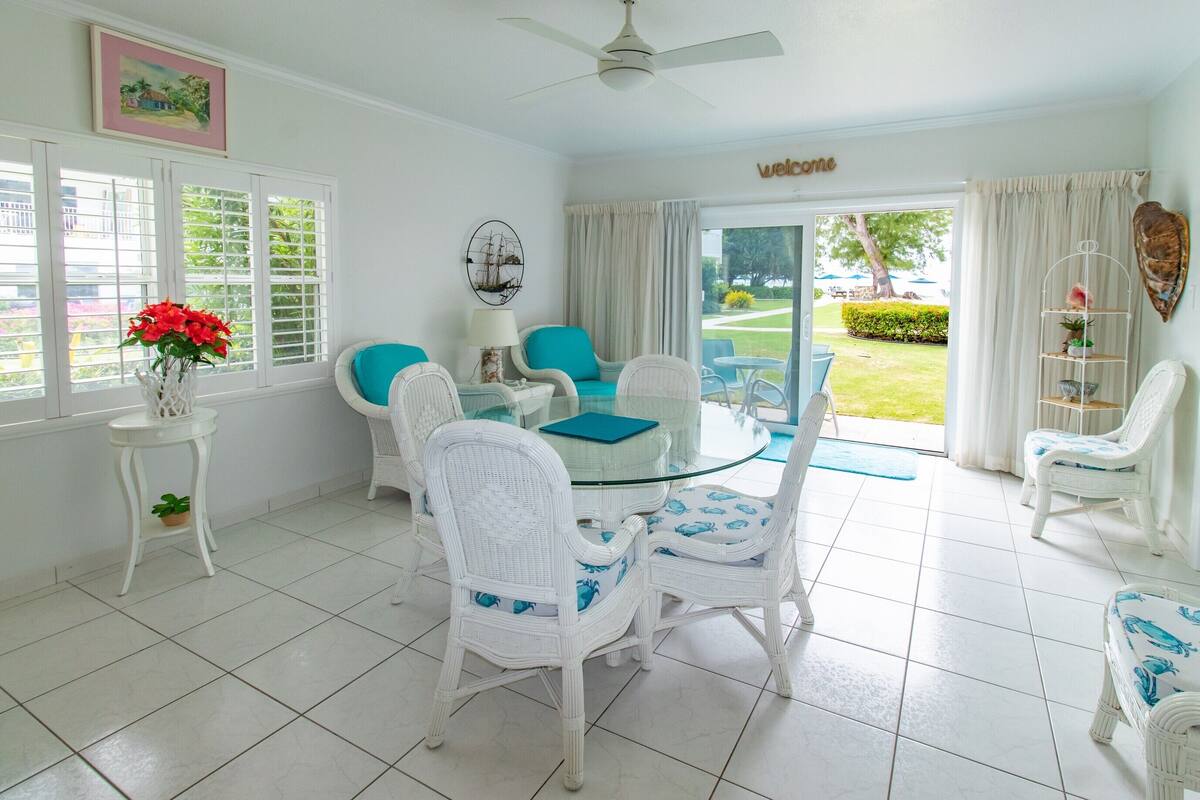 A bright room with white wicker furniture, a glass table, and a view of a garden through a sliding door.