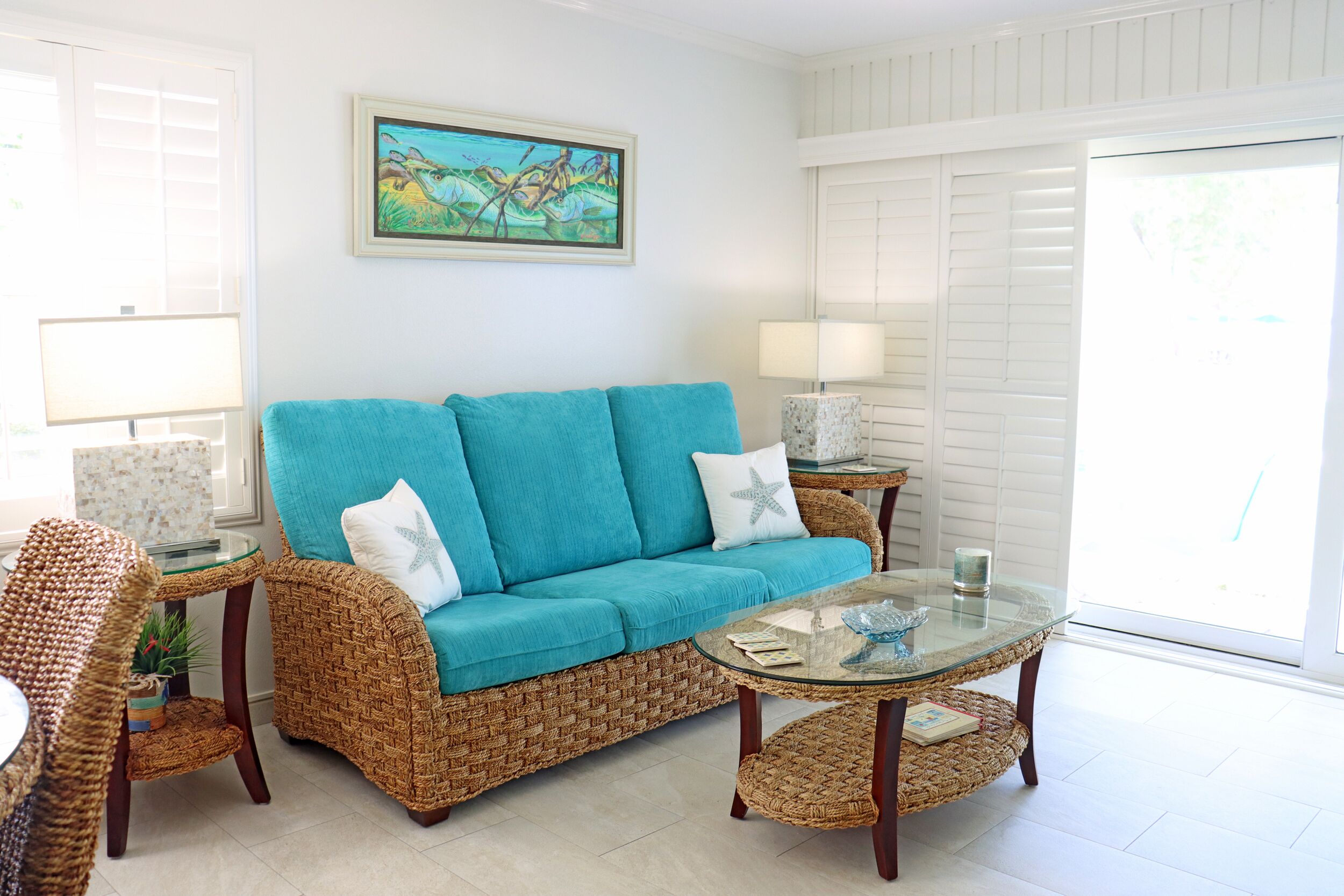 Un salon avec un canapé turquoise, des meubles en osier, une table basse en verre et un tableau accroché au mur.