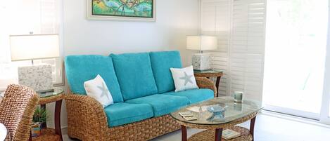 Un salon avec un canapé turquoise, des meubles en osier, une table basse en verre et un tableau accroché au mur.