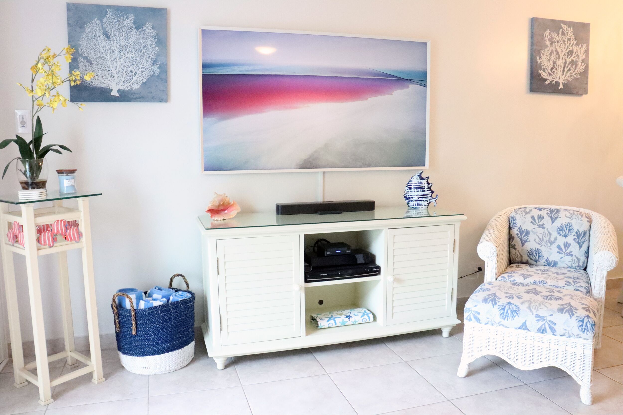 A living room with a white cabinet, a wicker chair, a glass table, and artwork on the wall.