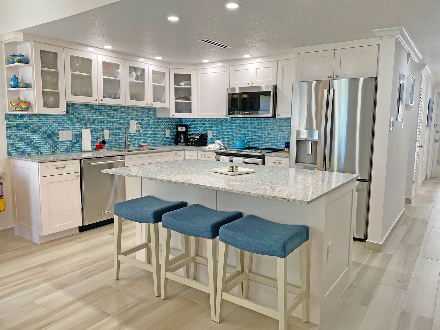 A modern kitchen with white cabinets, a blue-tiled backsplash, a stainless steel refrigerator, and a central island with bar stools.
