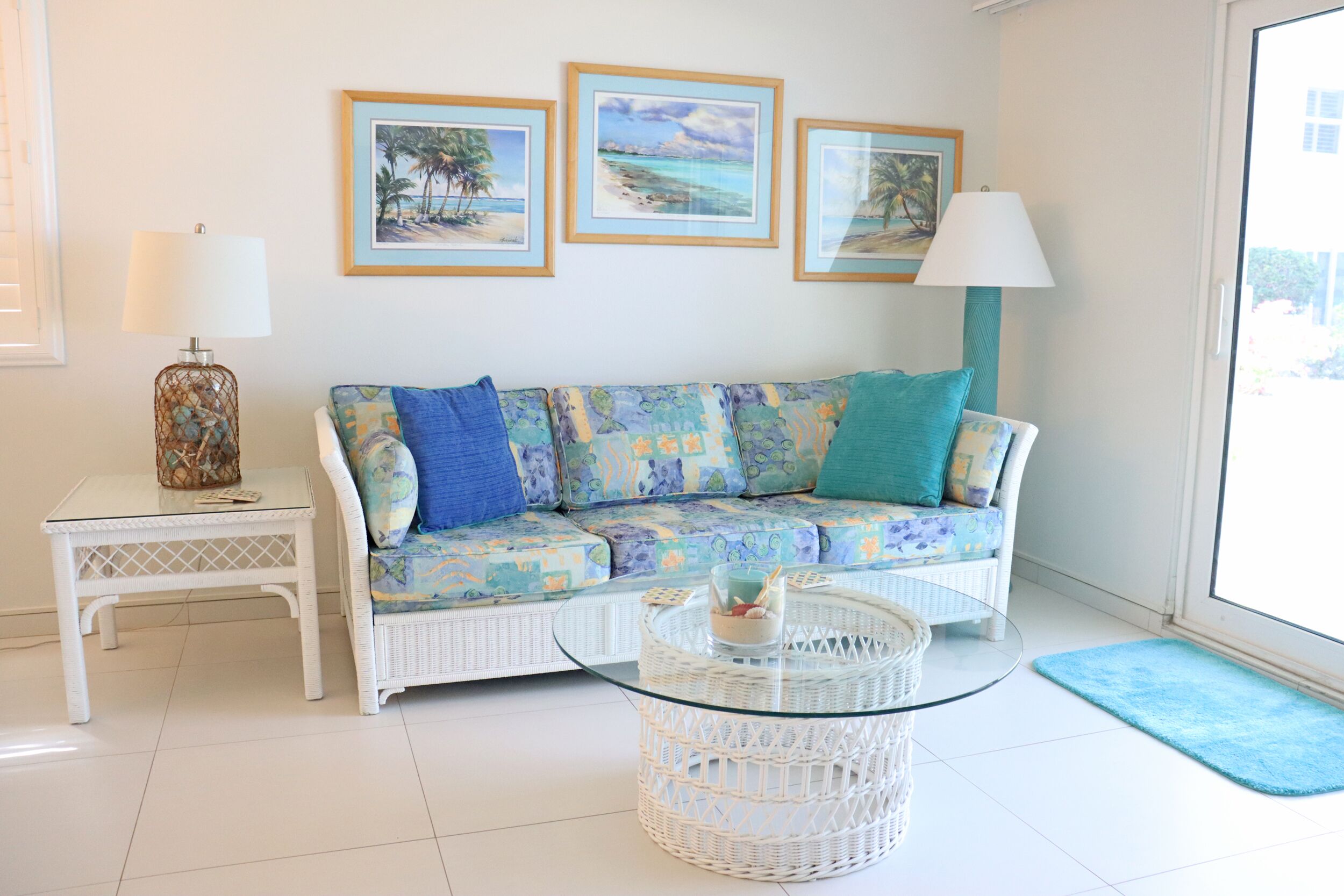 A living room with a white sofa, a glass coffee table, a wicker side table, and a door leading to a balcony.