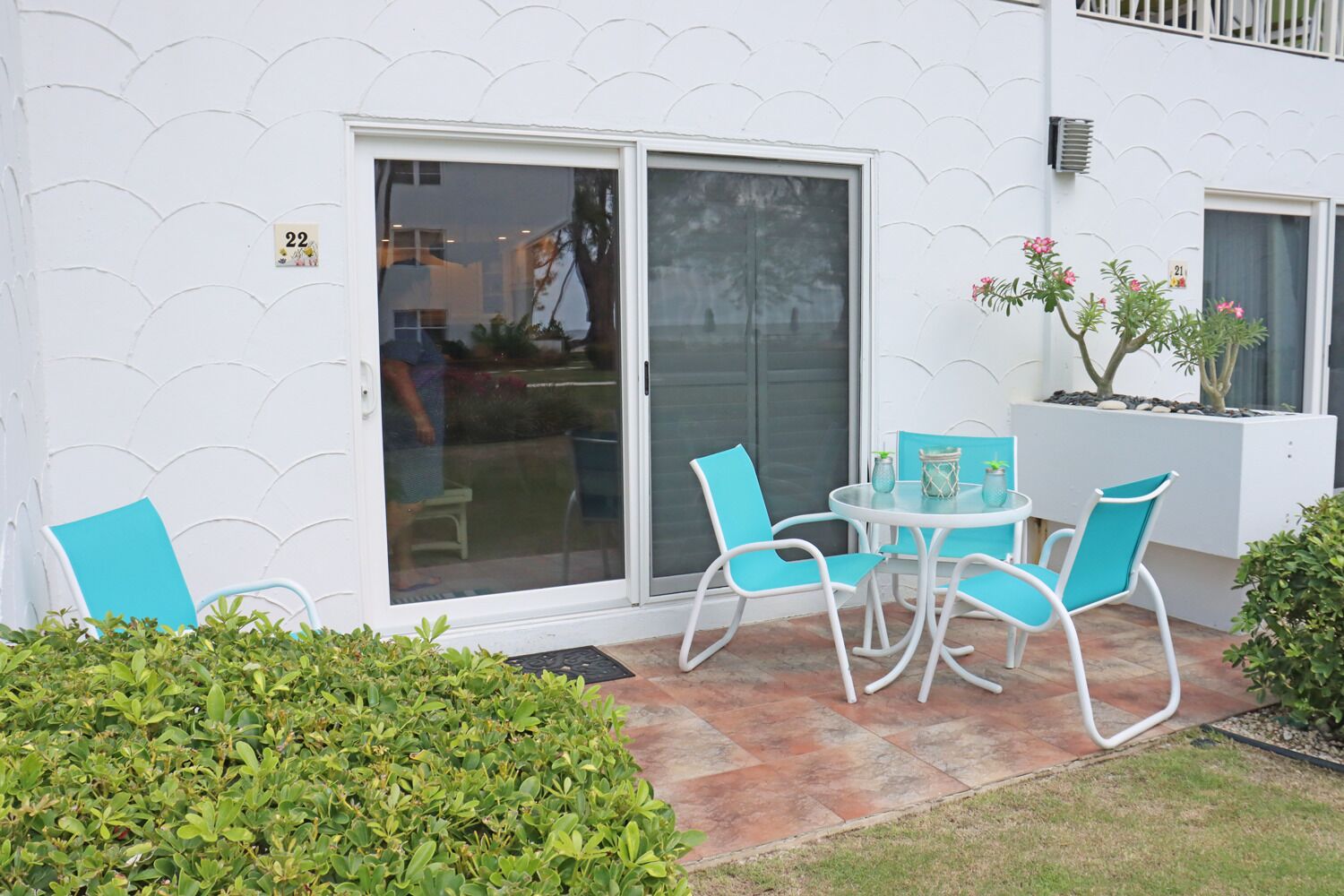 Un patio con pared blanca, puerta corredera de vidrio, una mesa pequeña con sillas y una planta en maceta.