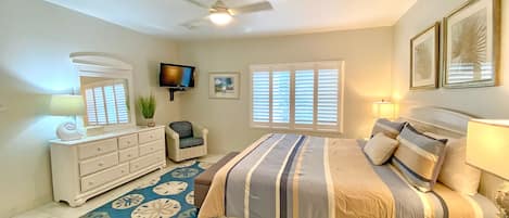 Un dormitorio con cama, cómoda, silla, televisor, ventilador de techo y ventanas.