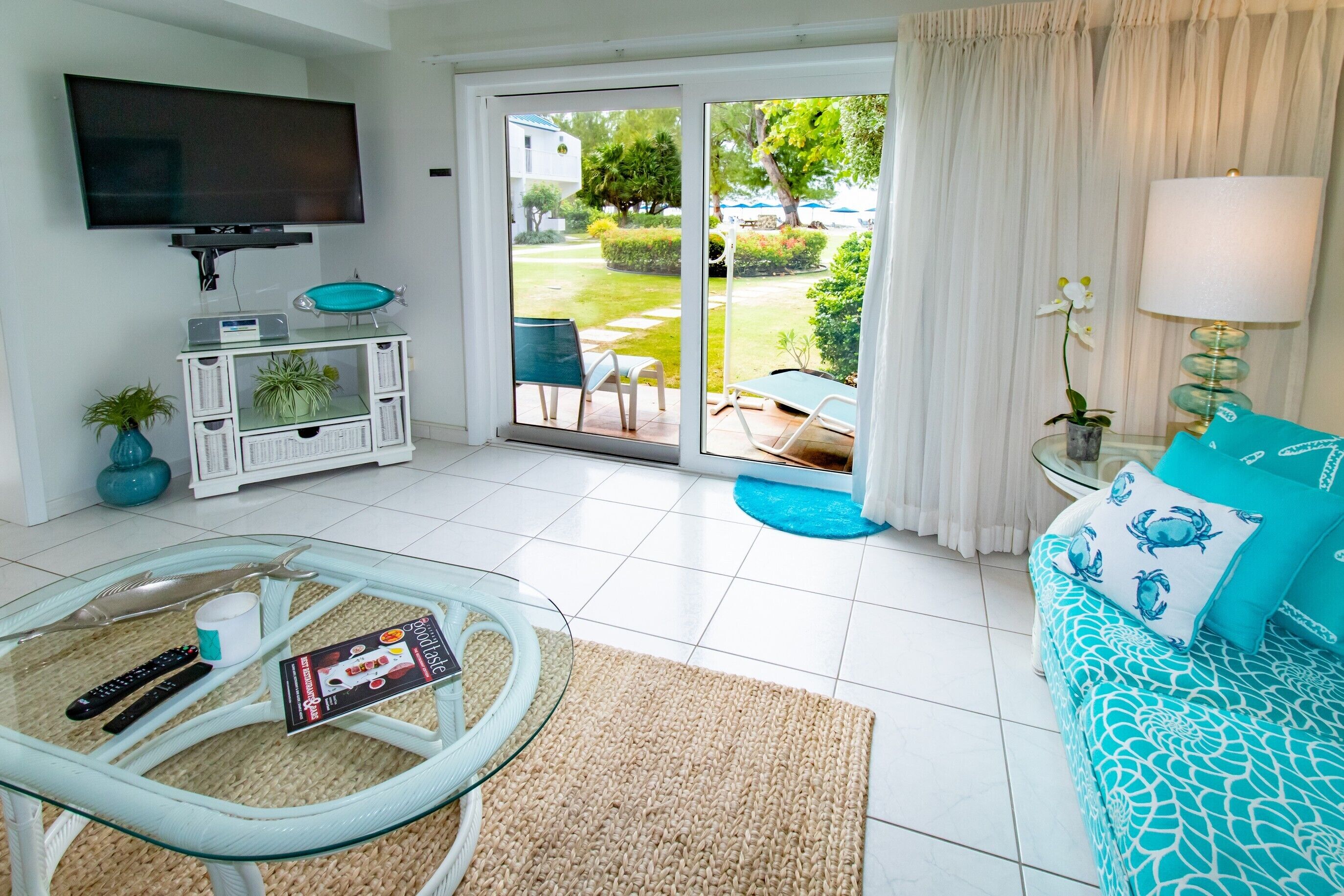 Ein modernes Wohnzimmer mit einem Glastisch, einer blauen Sofakissen-gepolsterten Couch, einem flachbildfernseher an der Wand und einer Glasschiebetür, die zu einem Garten führt.