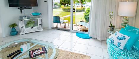 Ein modernes Wohnzimmer mit einem Glastisch, einer blauen Sofakissen-gepolsterten Couch, einem flachbildfernseher an der Wand und einer Glasschiebetür, die zu einem Garten führt.