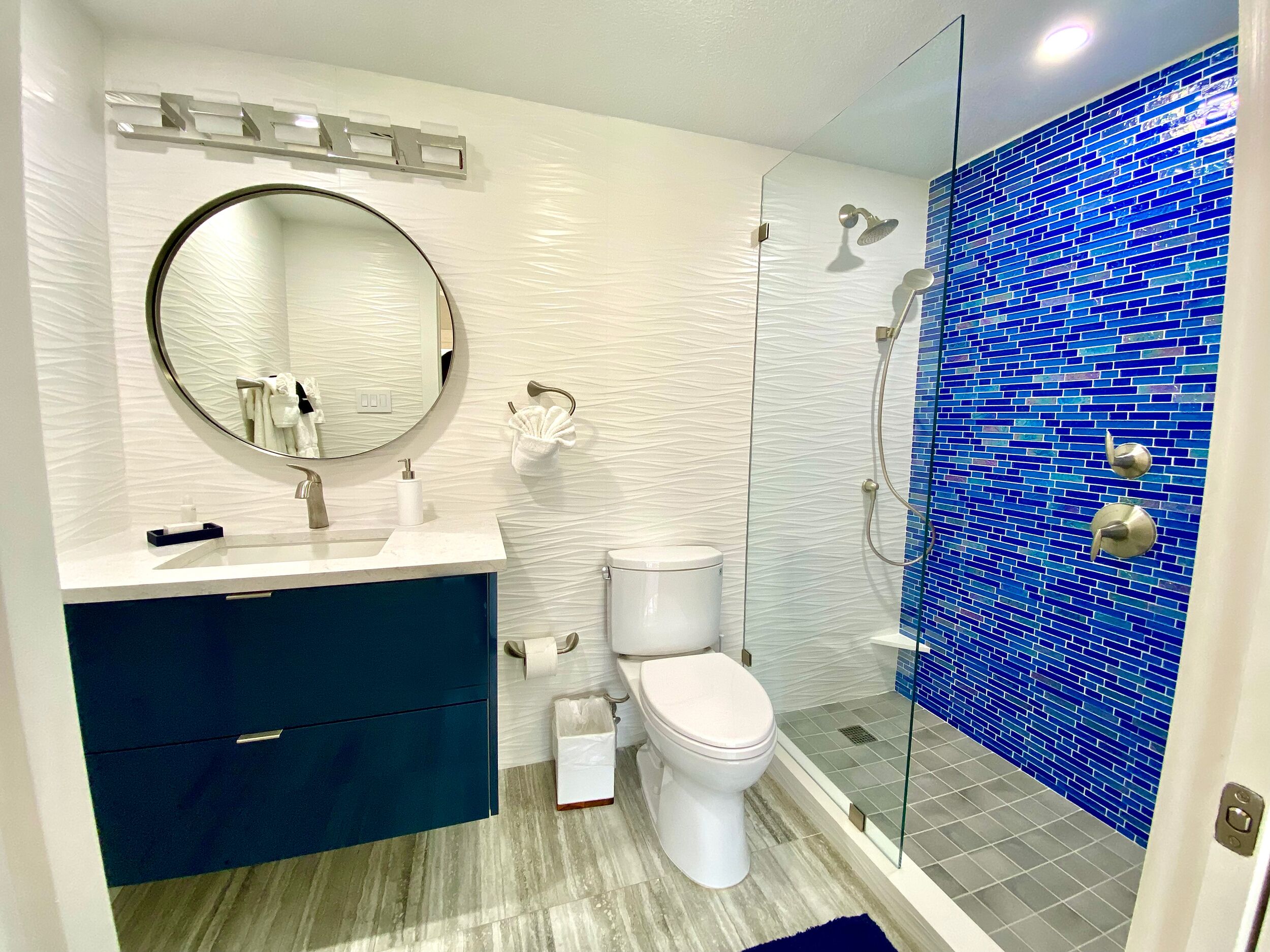 Ein modernes Badezimmer mit Toilette in Weiß, einem runden Spiegel, einer Duschkabine mit blauen Fliesen und einem dunkelblauen Waschtisch.