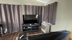 Sala de estar com televisão de tela plana, sofá, mesa de centro e uma mesinha com controle remoto.