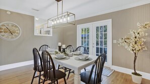Une salle à manger avec une table blanche dressée pour quatre personnes, entourée de chaises sombres. On peut voir une grande horloge murale et un miroir.
