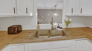 Une cuisine avec des meubles blancs, un évier en acier inoxydable, un plan de travail en bois, un grille-pain et des objets de décoration.
