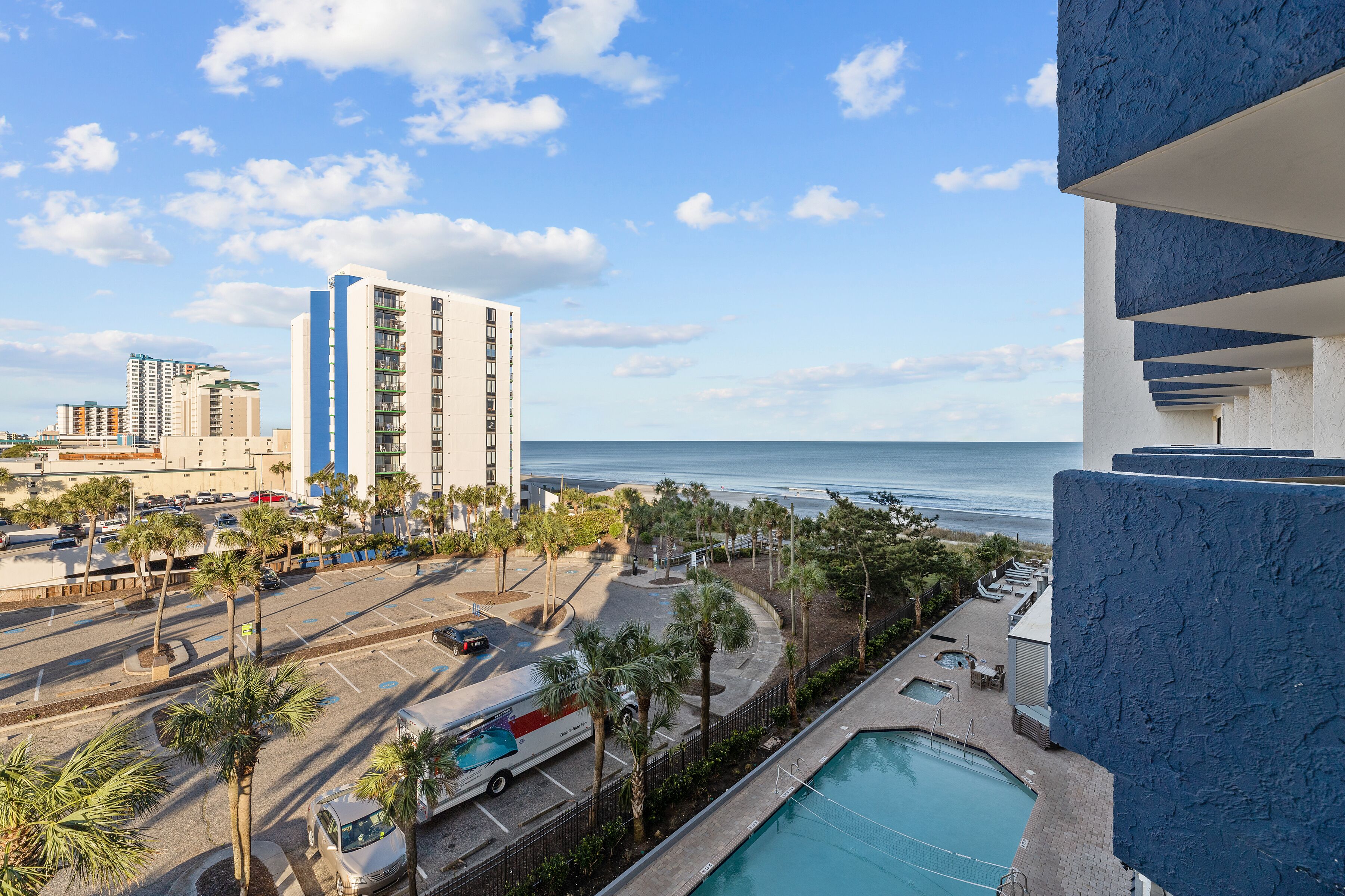 A high-rise hotel with a pool, a view of the beach, and a parking area with palm trees.