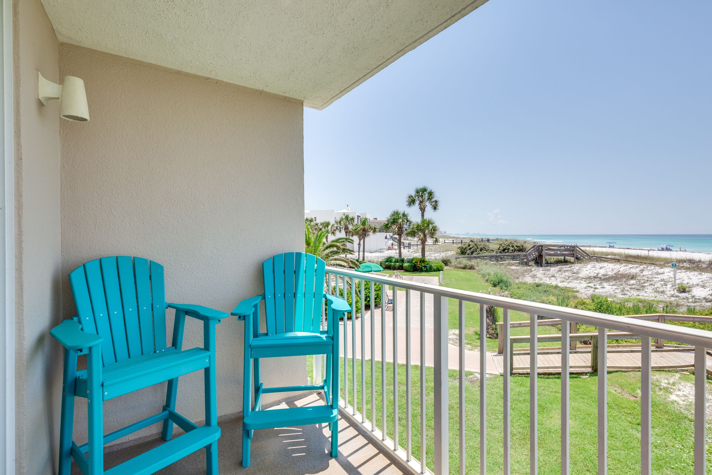 Un balcon avec deux chaises turquoise, une balustrade blanche et une vue sur la plage et l’océan.