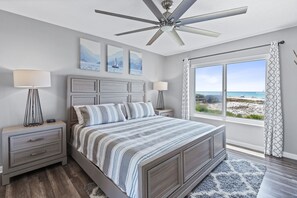 A bedroom with a bed, two bedside tables, a ceiling fan, and a large window with a view of the beach.
