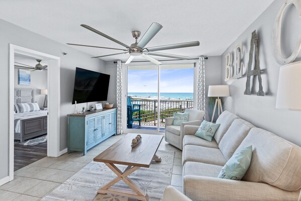 A modern living room with a ceiling fan, a TV, a sofa, and a view of the beach.