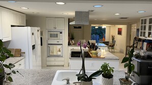 A modern kitchen with white cabinets, a built-in microwave, oven, and sink. There are potted plants on the counter and a dining area visible in the background. - House on water canal, 5 minutes from downtown and beach. (Naples)