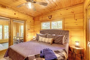 Un dormitorio tipo cabaña de madera con una cama grande, dos ventanas, un ventilador de techo y una pequeña mesa con un farol.
