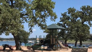 A playground with a slide, swings, and a covered play area, surrounded by trees and benches. - Lake Travis townhome-Pool-Marina-Boat ramp-Golf (Point Venture)
