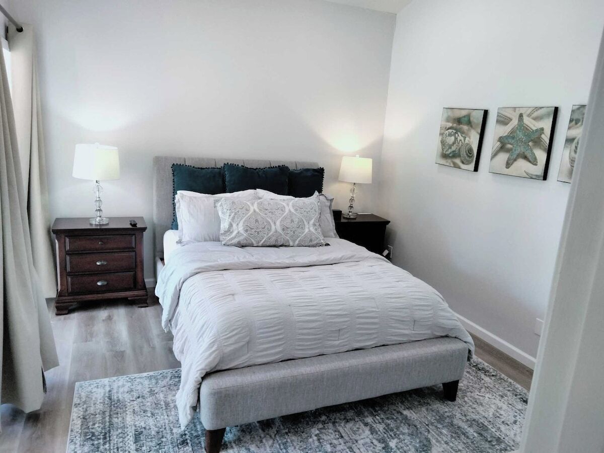 A bedroom with a bed, two bedside tables, a nightstand, a patterned rug, and wall art.