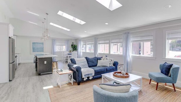 A modern living room with a sofa, coffee table, and chairs. There is a kitchen area with a refrigerator and cabinets. The room has large windows with curtains.