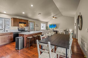 Une cuisine spacieuse avec des meubles en bois, un îlot central doté de tabourets, un coin repas avec une longue table et des chaises, et un ventilateur de plafond.