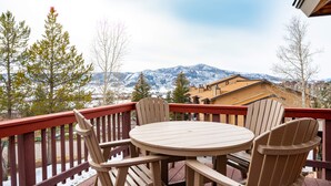 Una terraza de madera con una mesa redonda y cuatro sillas, con vistas a una cadena montañosa nevada.