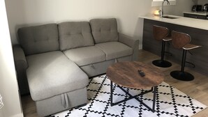 A modern living room with a grey sofa, a wooden coffee table, and two bar stools. A kitchen area with a microwave and toaster is visible in the background. - A - Modern Cozy Room w Private Entrance & Washroom (Surrey)