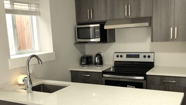 A modern kitchen with dark wood cabinets, a stainless steel microwave, a built-in oven, and a large island with bar stools. - A - Modern Cozy Room w Private Entrance & Washroom (Surrey)