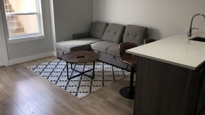 A compact living space with a sofa, a small round table, a kitchen area with a sink, and a patterned rug on the floor. - A - Modern Cozy Room w Private Entrance & Washroom (Surrey)