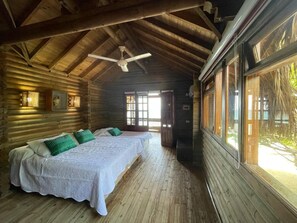 Una habitación tipo cabaña de madera con dos camas, un ventilador de techo y ventanas grandes.