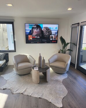 A modern living room with a flat-screen TV mounted on the wall, two beige armchairs, a glass coffee table, and a large potted plant.