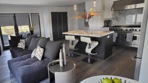 Eine moderne Küche mit einer zentralen Kochinsel, ein Essbereich mit Barhocker und ein Wohnzimmer mit einem dunkelgrauen Sofa.