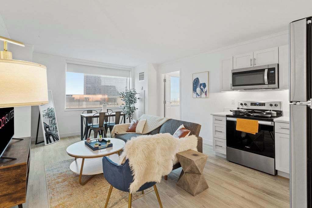 A modern kitchen with stainless steel appliances, a dining area with a round table and chairs, and a living room with a sofa and a TV.