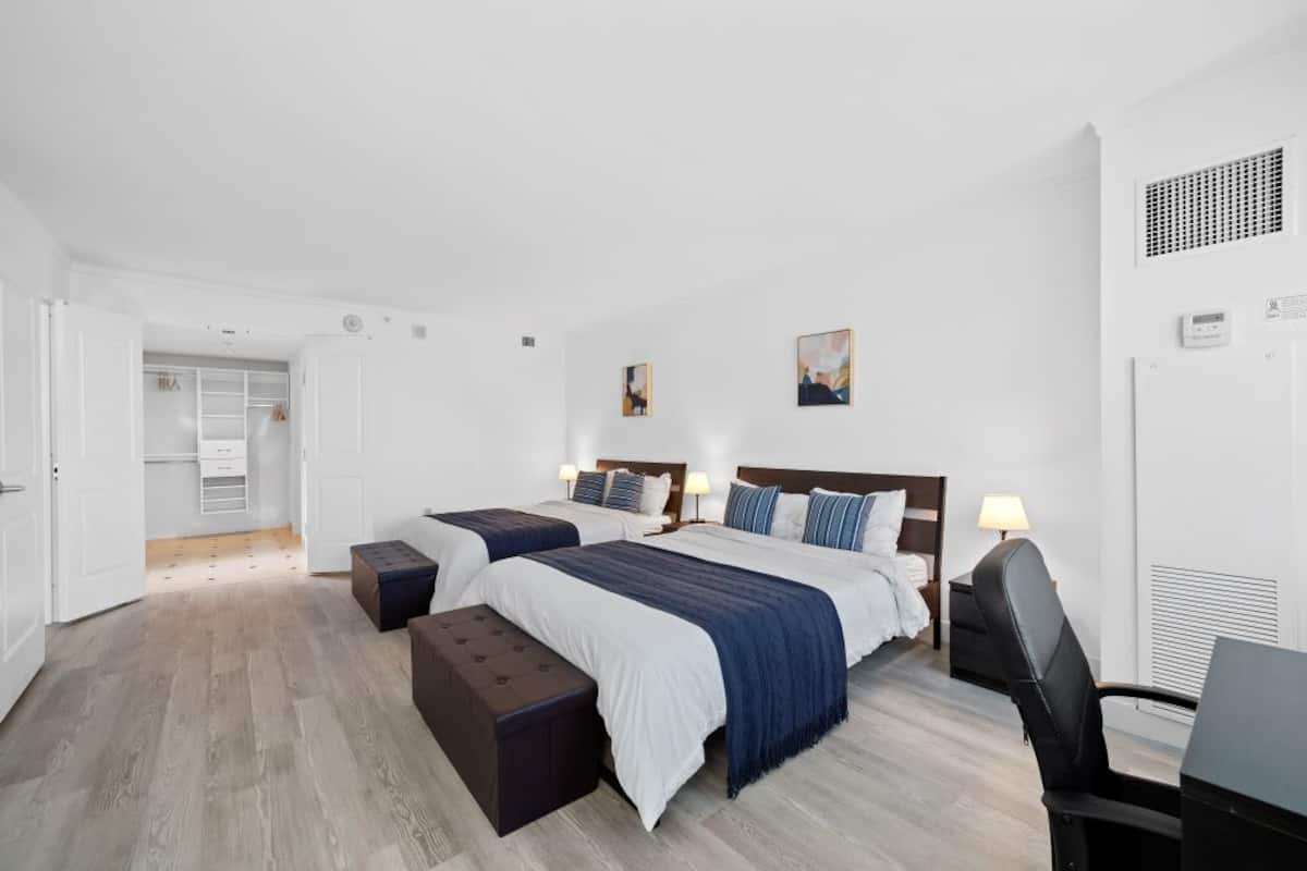 A hotel room with two beds, a desk, and a chair. There is a wardrobe and a small table with a lamp.