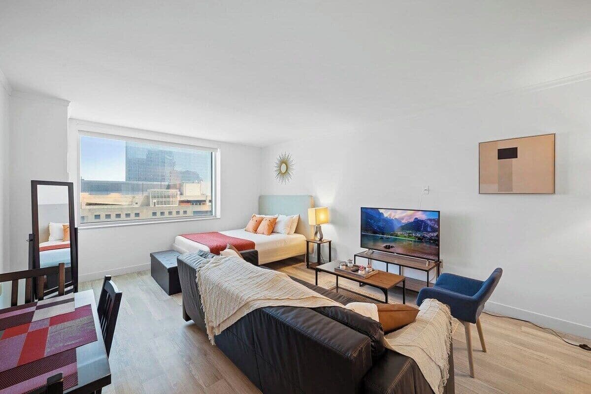 A modern hotel room with a large window, a bed, a sofa, a TV, and a dining area.