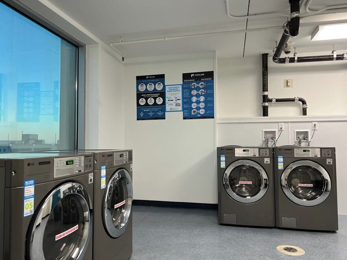 A laundry facility with two front-loading washers and dryers, a window with a view of buildings, and informational posters on the wall.