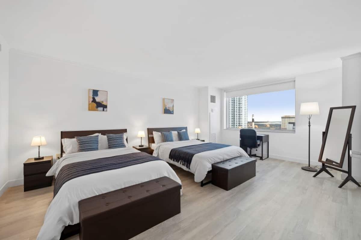 A hotel room with two beds, a desk, a chair, a standing lamp, and a window with a city view.