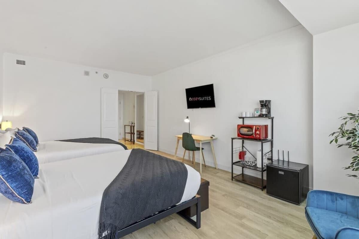 A bedroom with a bed, a desk, a TV, a coffee machine, and a small fridge.