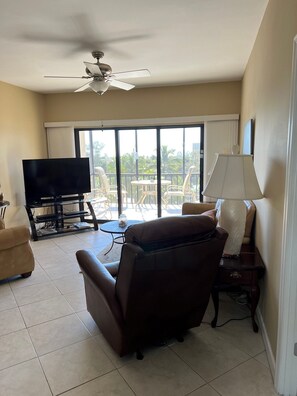 A living room with a TV, a ceiling fan, a sofa, a table, and a lamp. - Penthouse Condo with Ocean View in the Heart of Siesta Key (Sarasota)