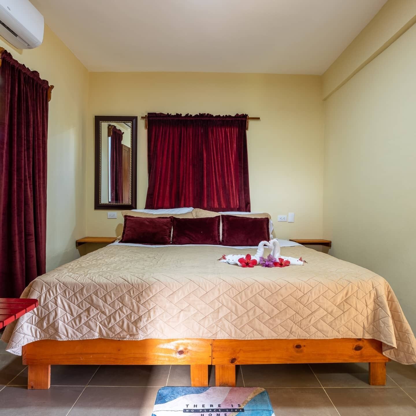 Una habitación de hotel con una cama, cabecera de madera, mesita de noche, espejo y cortinas rojas.