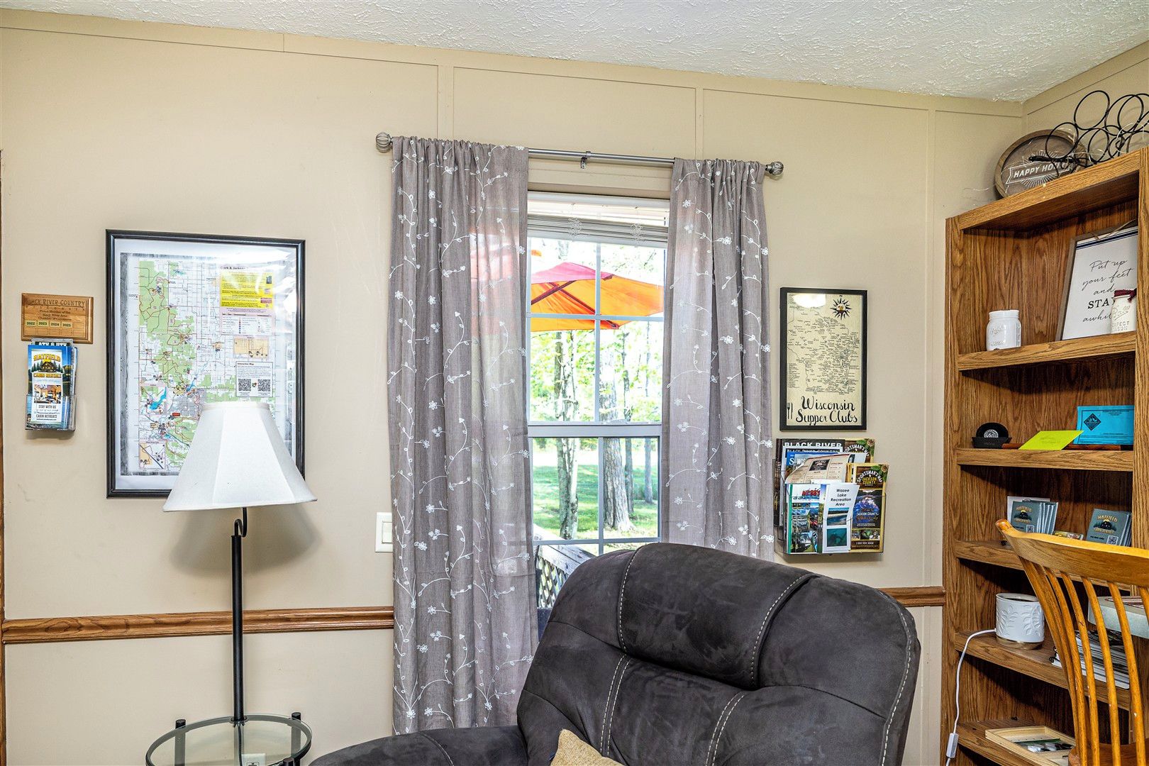 A room with a large window, a map on the wall, a lamp, a chair, and a bookshelf with various items.