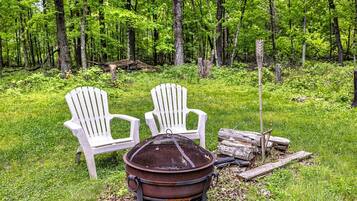 Two Adirondack chairs, a metal fire pit, and a log in a forested area.