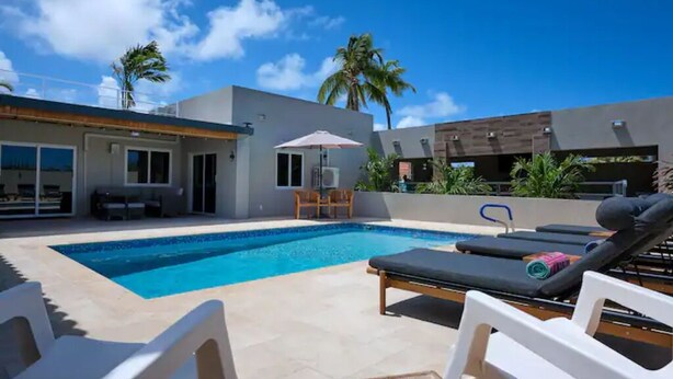 A modern outdoor pool area with lounge chairs, a table, and a covered seating area. - Beautiful fully gated private home with resort like amenities (Noord)