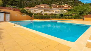 A swimming pool with a clear blue water, surrounded by a tiled deck. In the background, there is a hillside with buildings and greenery. - Nice apartment for 4 people with WIFI, pool, TV and terrace (Le Lavandou)