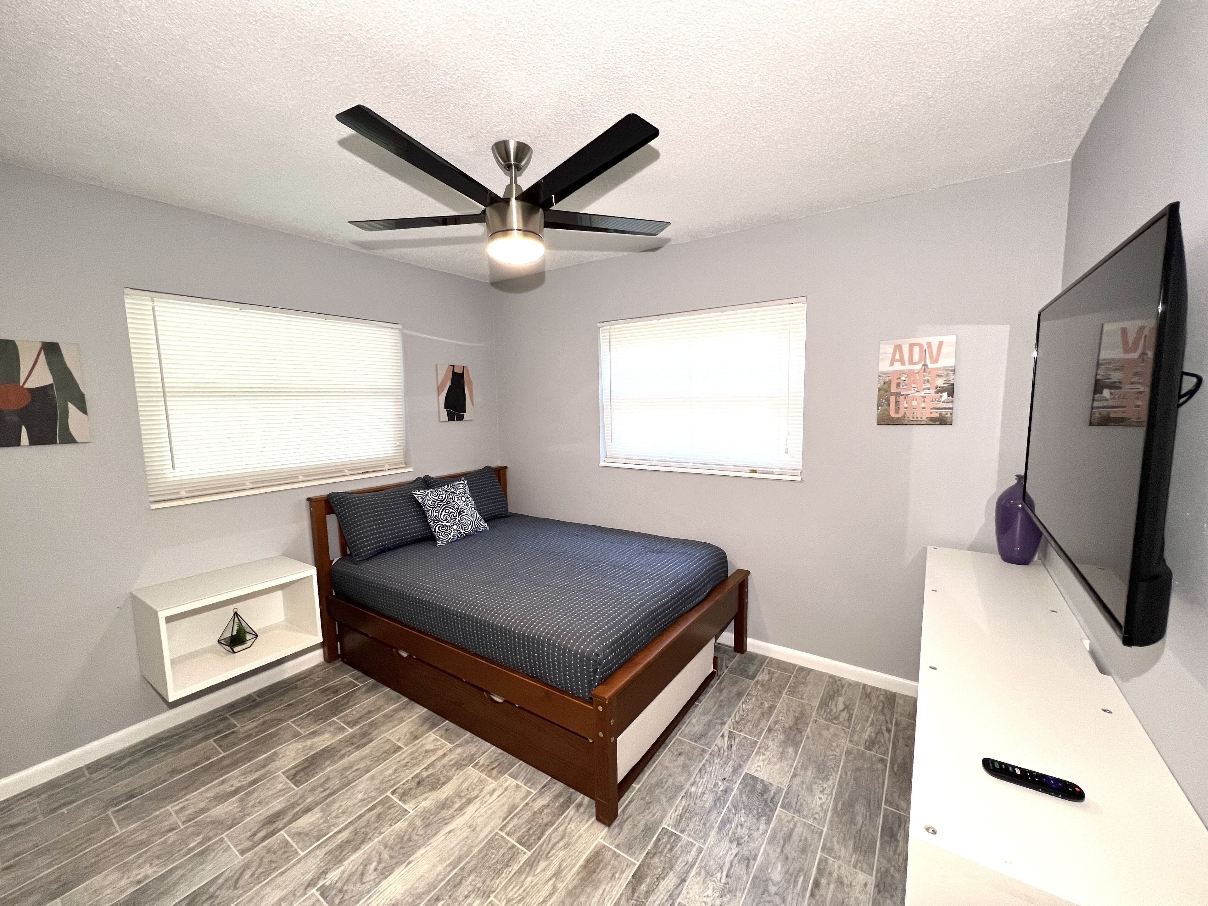A bedroom with a bed, a ceiling fan, a TV, and a white nightstand.