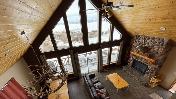 A cozy cabin interior with a stone fireplace, leather sofas, a wooden dining table, and large windows offering a view of a snowy landscape.