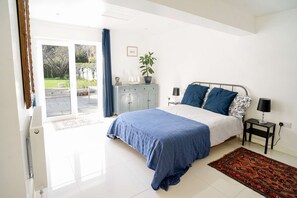 A bedroom with a bed, a nightstand, a lamp, a potted plant, and a view of the outdoors.