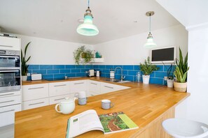 A modern kitchen with white cabinets, blue backsplash, wooden countertop, hanging pendant lights, and potted plants.